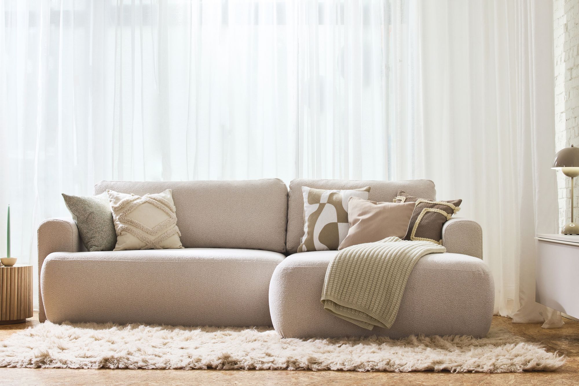 Hellbeiges Sofa mit abgerundeten Armlehnen, dekorativen Kissen und einer grünen Decke auf einem Flokati-Teppich